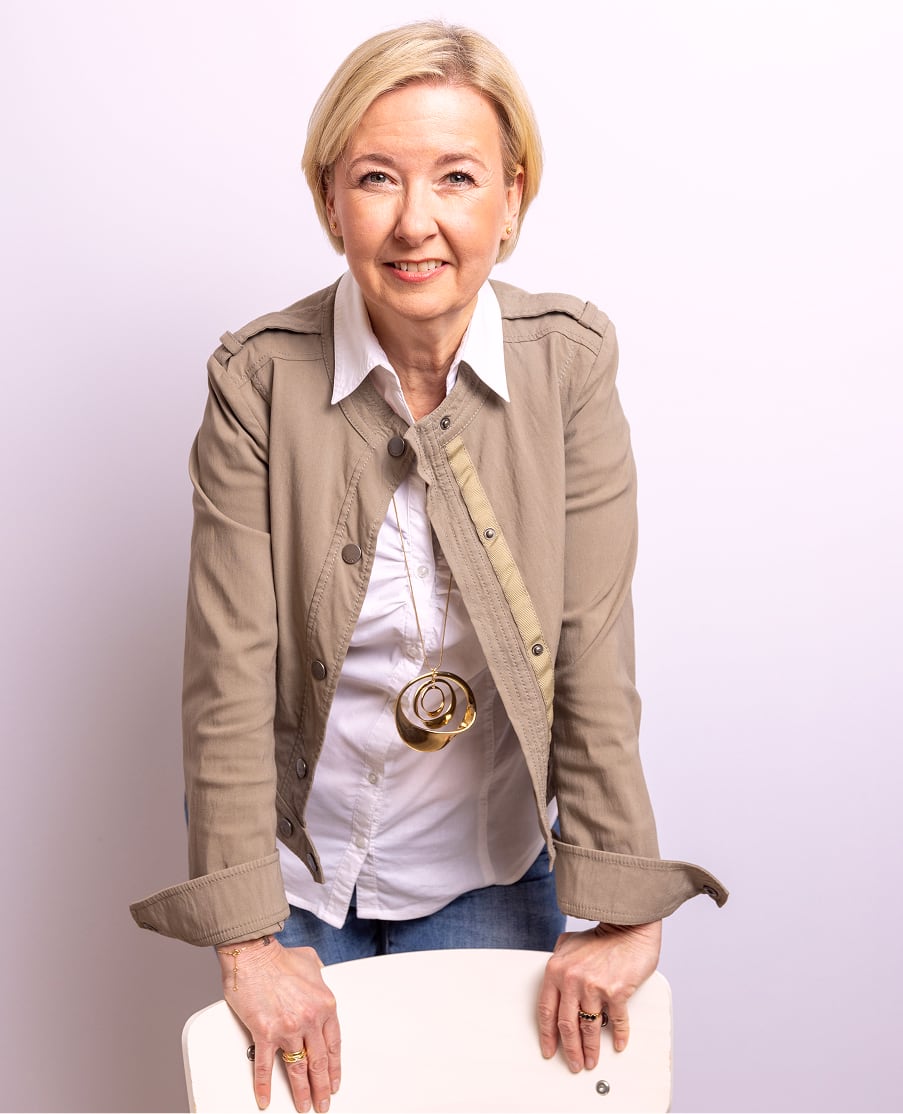 Angela Schuh-Haunold in beiger Jacke und weißer Bluse, lächelnd auf einen Stuhl gestützt